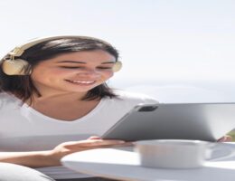 Pessoa relaxando em ambiente externo enquanto utiliza um tablet e escuta música com fones grandes, ao lado de uma xícara de café